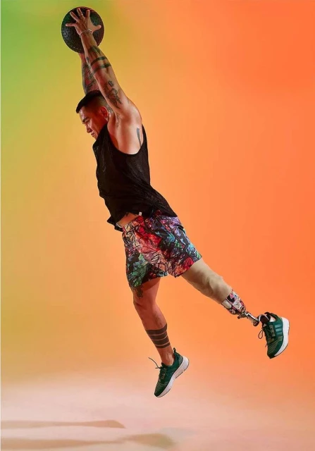Athletic man with prosthetic leg leaps mid-air catching a medicine ball, illuminated by studio lighting against an orange gradient backdrop.