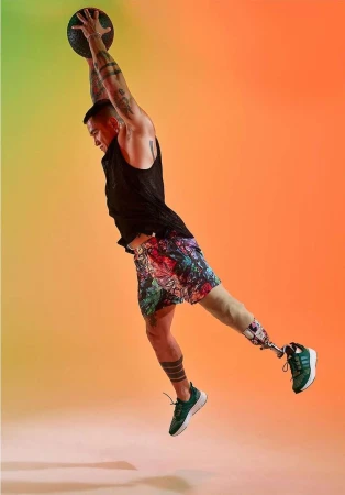 Athletic man with prosthetic leg leaps mid-air catching a medicine ball, illuminated by studio lighting against an orange gradient backdrop.