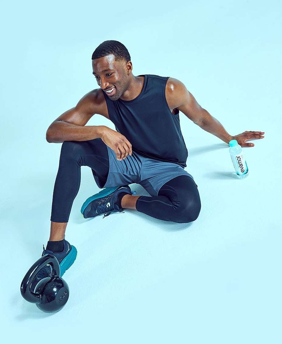 Athletic Black man in activewear sits on a blue backdrop with a kettlebell and water bottle, smiling during a fitness workout session.