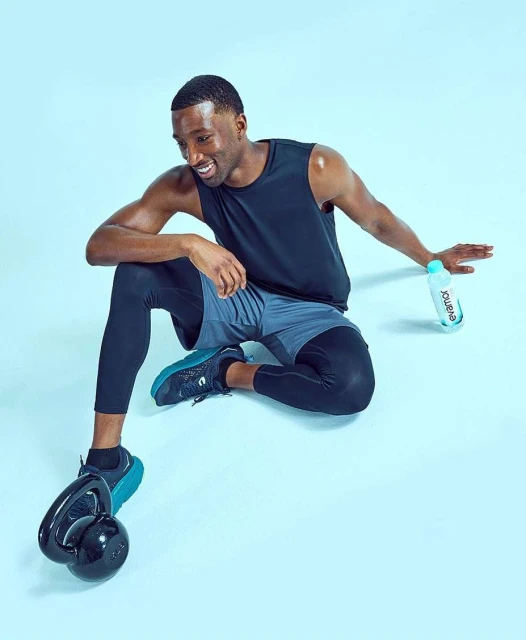 Athletic Black man in activewear sits on a blue backdrop with a kettlebell and water bottle, smiling during a fitness workout session.