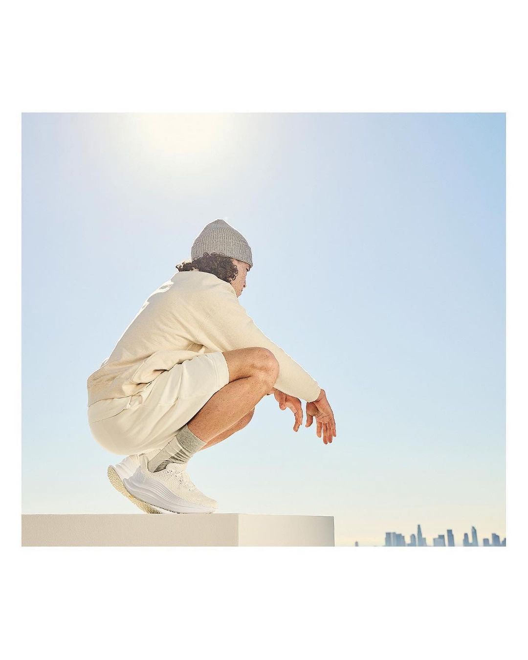 Man wearing a beanie, shorts, and shoes crouches on a white block against a blue sky with a city skyline in the distance.