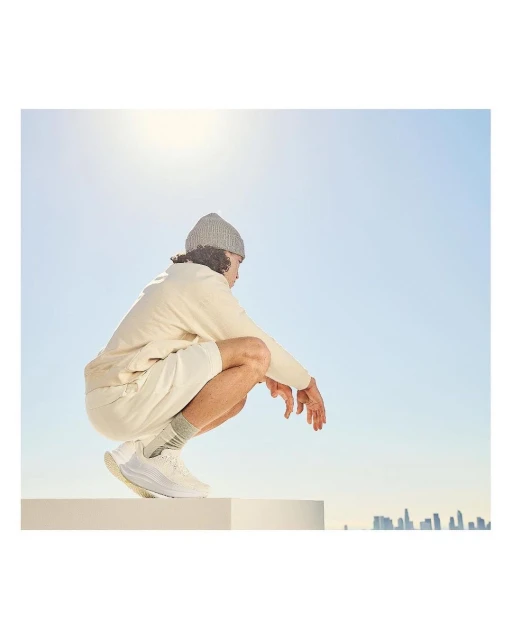 Man wearing a beanie, shorts, and shoes crouches on a white block against a blue sky with a city skyline in the distance.