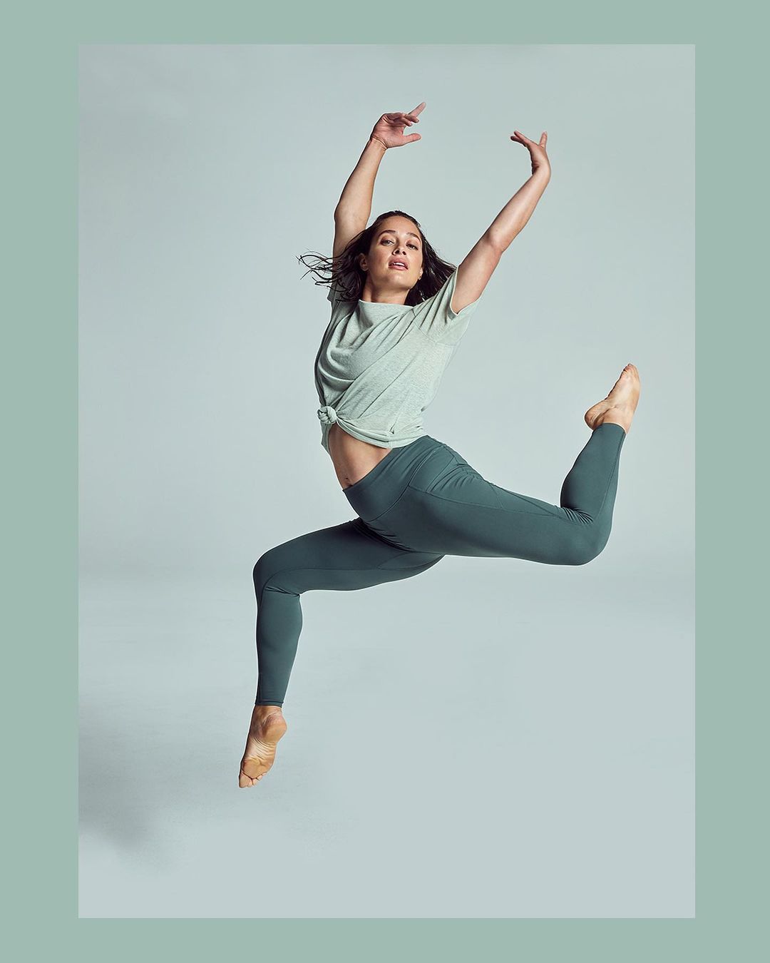 A dancer mid-air wearing a light green top and dark green leggings against a light gray backdrop in a dance pose.