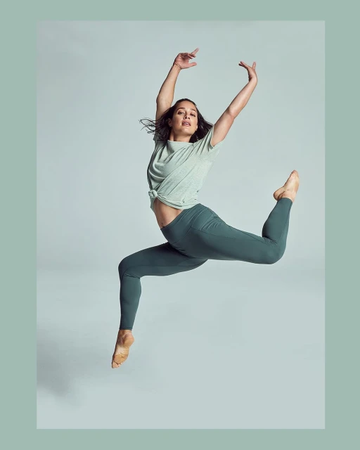 A dancer mid-air wearing a light green top and dark green leggings against a light gray backdrop in a dance pose.