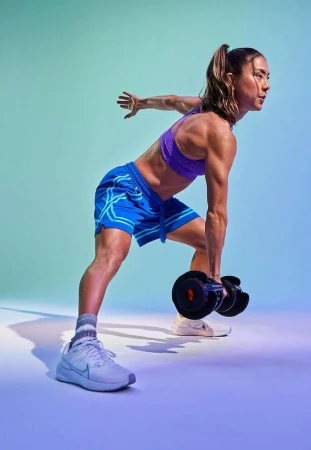 Athletic woman in sports bra and shorts performs a single-arm dumbbell row in a fitness studio with teal and purple lighting.
