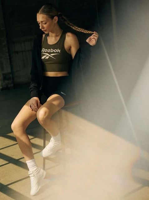 Athletic woman in Reebok sports bra and shorts, sitting and holding her braid, captured in an indoor setting with soft, diffused light.