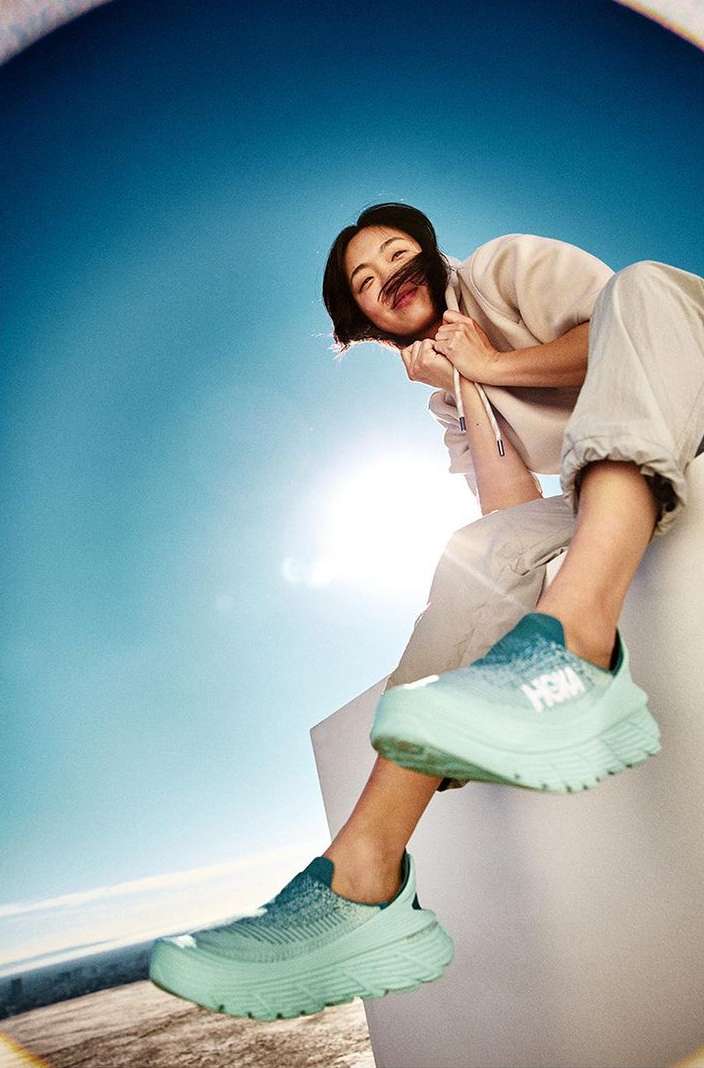 Eye-level view of an Asian woman sitting atop a white structure wearing light-green Hoka sneakers, against a bright-blue sky.
