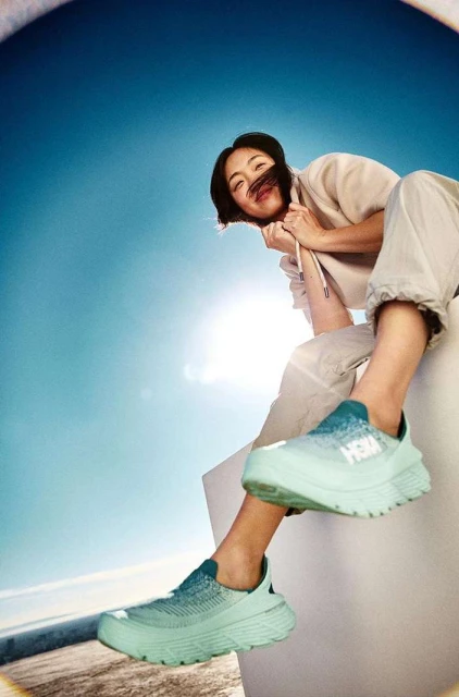 Eye-level view of an Asian woman sitting atop a white structure wearing light-green Hoka sneakers, against a bright-blue sky.