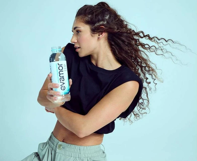 Adult woman in a studio shot, holding an Evamor water bottle. She is wearing a dark crop top and light-colored pants with loose curly hair.