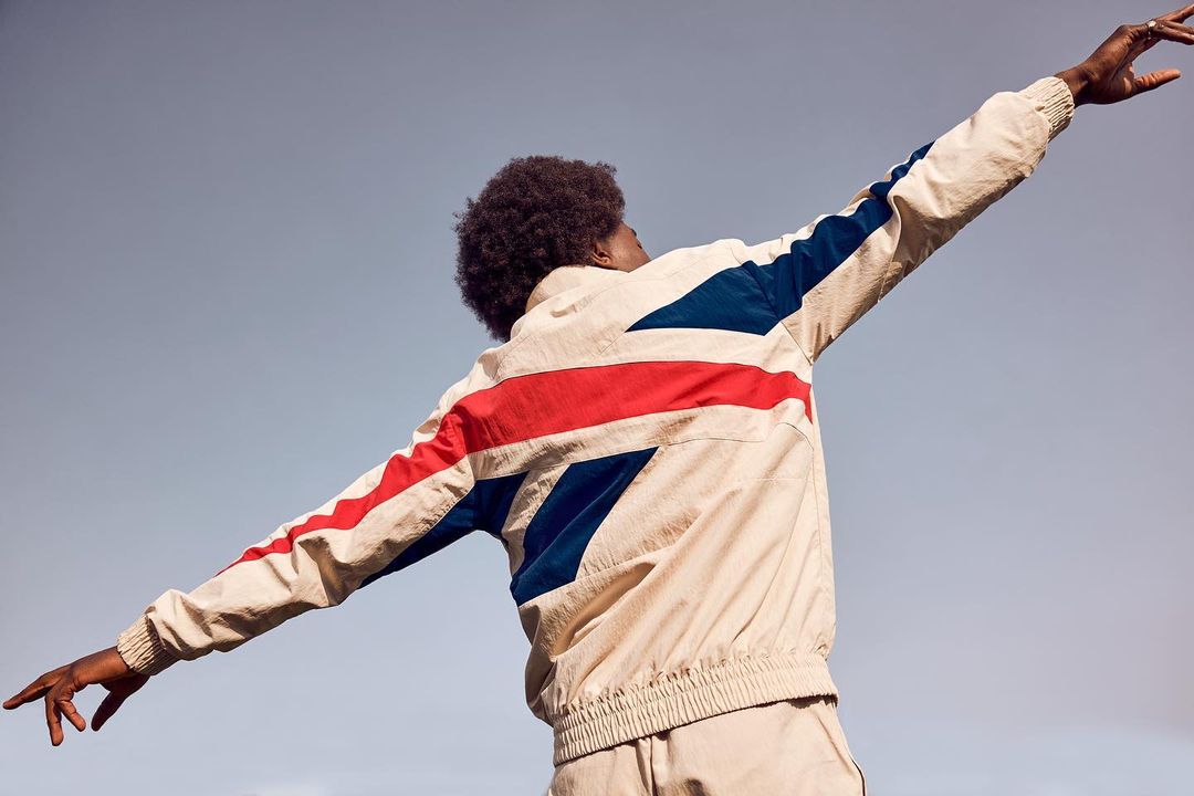 An adult with dark hair wears a cream and red and blue jacket with the Union Jack flag, arms outstretched, photographed from a low angle.