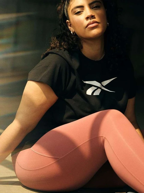 Adult female wearing a black Reebok t-shirt and pink leggings is posing in a medium shot, with sunlight coming from the side.