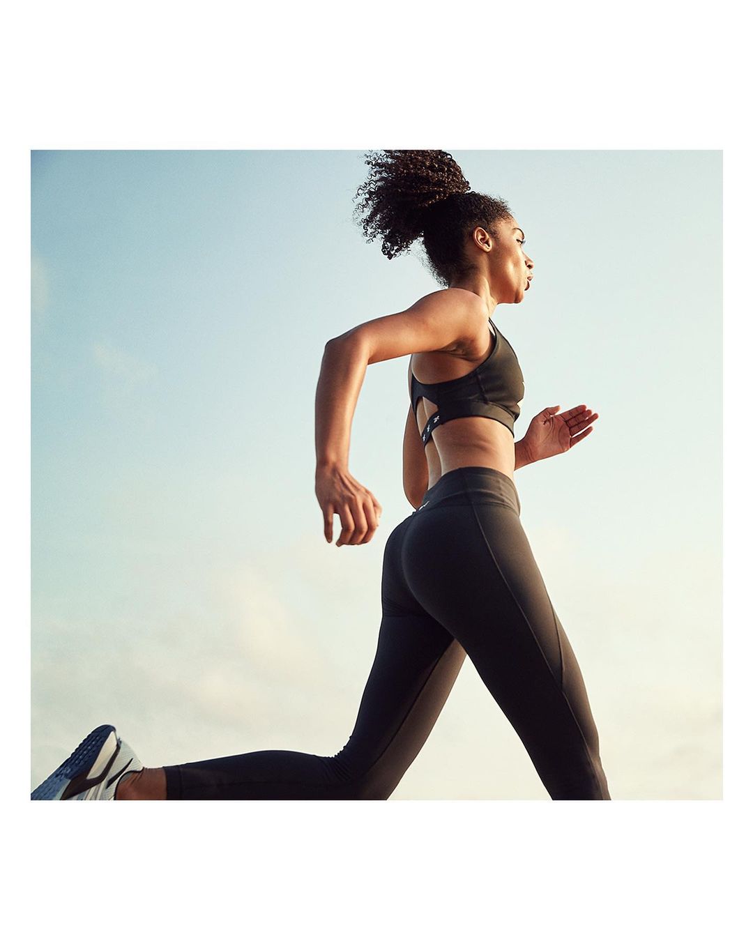 An athletic African-American woman runs in black leggings and a sports bra against a blue sky, showcasing fitness and determination.