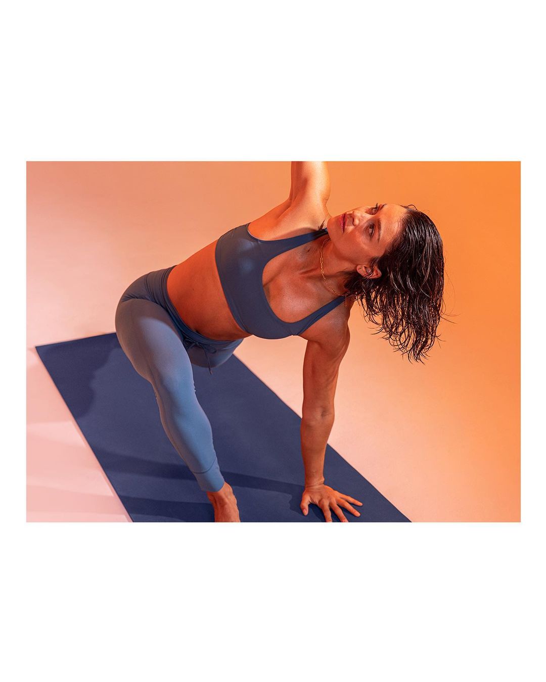 A woman in sportswear executes a yoga pose on a blue mat against an orange background, captured in a high-angle medium shot.