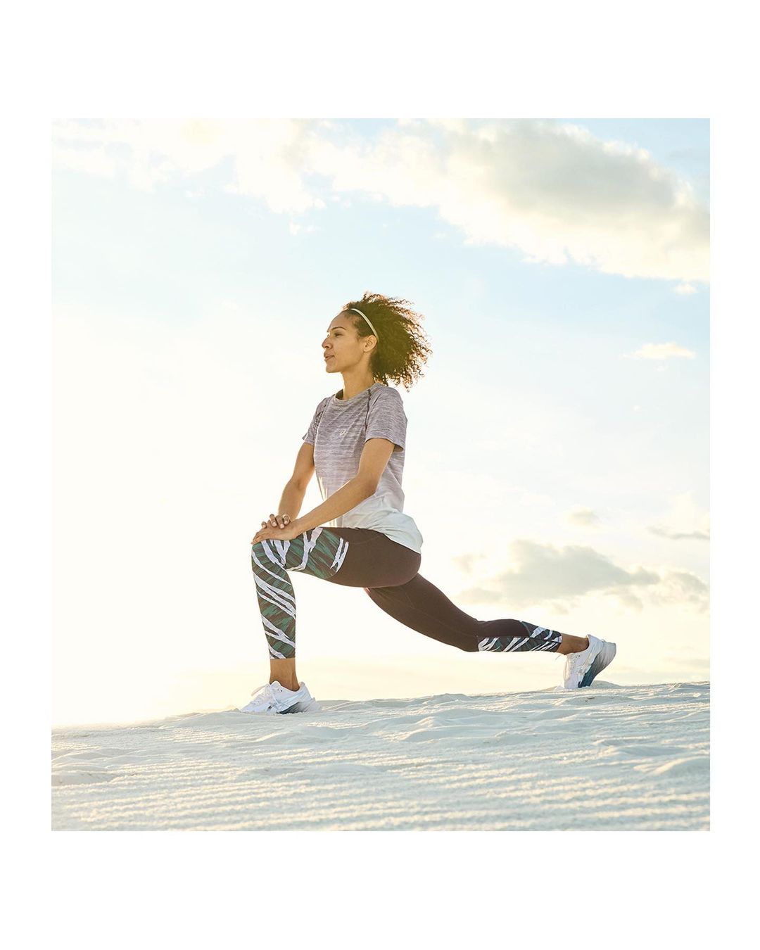 Athletic adult woman stretches her leg muscles outdoors under a bright, cloudy sky for a fitness workout. She wears athletic leggings and top.