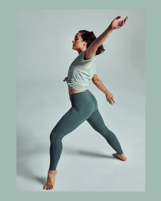 A woman with brown hair is posed in an artistic pose with her arms extended in a photo studio wearing exercise clothing.