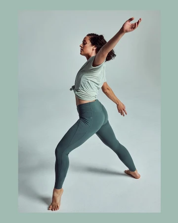 A woman with brown hair is posed in an artistic pose with her arms extended in a photo studio wearing exercise clothing.