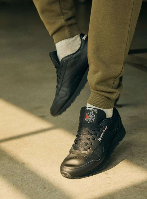 Close-up of person wearing olive-green sweatpants and grey socks, showcasing a pair of black Reebok Classic Leather sneakers with logos.