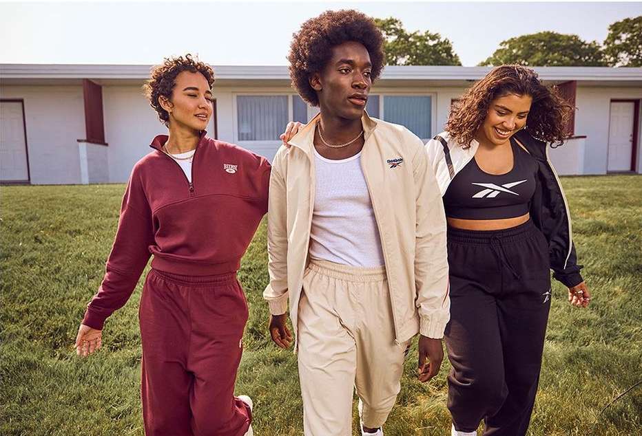 Three people in athletic wear walking on grass in front of a building. The models are wearing matching Reebok tracksuits.