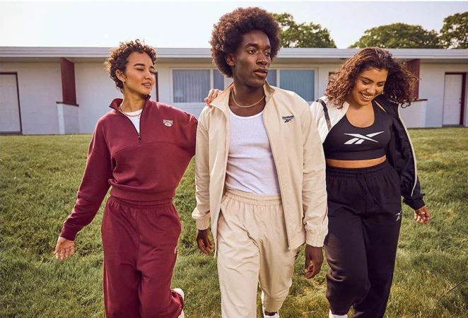 Three people in athletic wear walking on grass in front of a building. The models are wearing matching Reebok tracksuits.