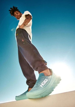 Eye-level view of a Black man in sweatpants and a t-shirt posing outdoors in turquoise slip-on sneakers with the Hoka logo, against a blue sky.