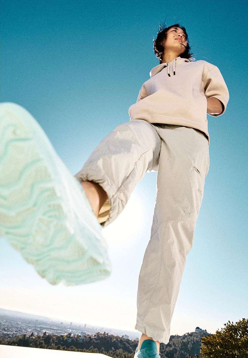 Low-angle shot of a young adult wearing a hoodie and neutral-toned pants, standing on a hill with cityscape in the background on sunny day.
