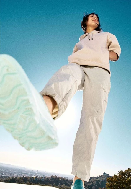 Low-angle shot of a young adult wearing a hoodie and neutral-toned pants, standing on a hill with cityscape in the background on sunny day.