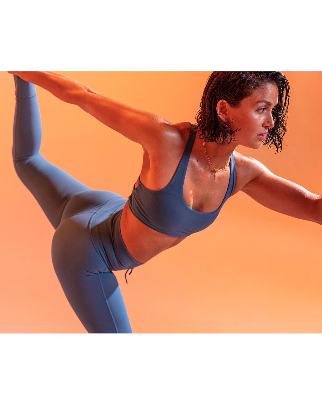 Athletic woman in blue workout attire performs a standing bow yoga pose against a peach background with studio lighting. 