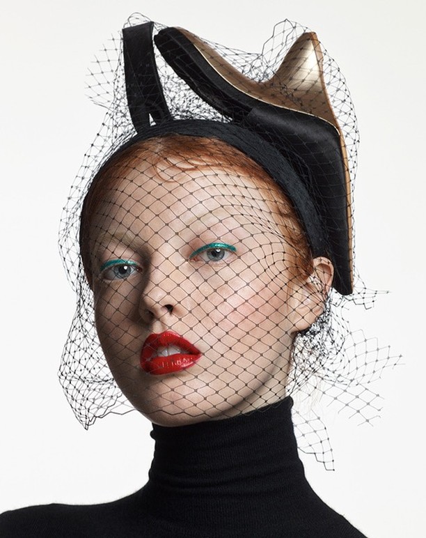Portrait of a woman wearing a black turtleneck and a black headpiece with a decorative gold high heel and netting against white background.