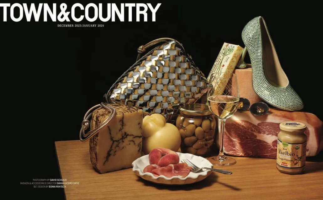 Still life editorial shot featuring a handbag, shoe, and gourmet foods. The image showcases luxury products in a dark studio.