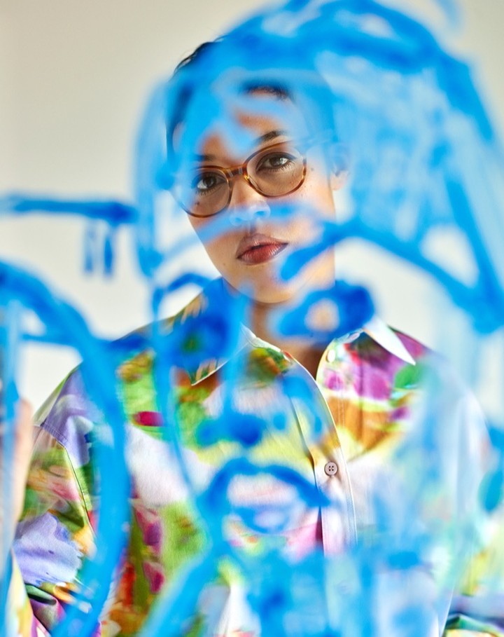 Close-up of a young woman with short hair and glasses, wearing a patterned blouse, standing in front of a blue painted design.