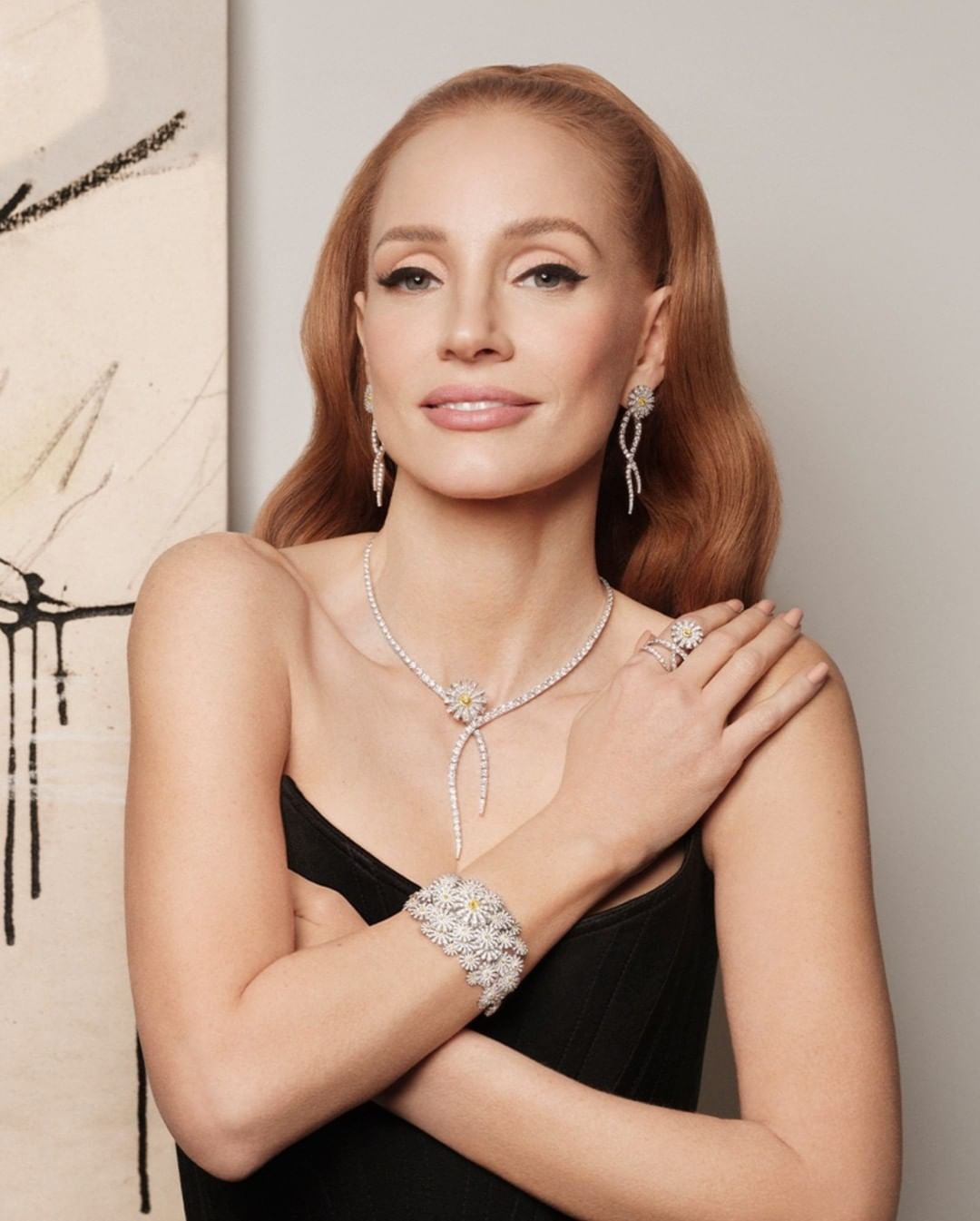Portrait of a redhead adult female wearing diamond jewelry, including a necklace, earrings, ring, and bracelet, posing indoors.
