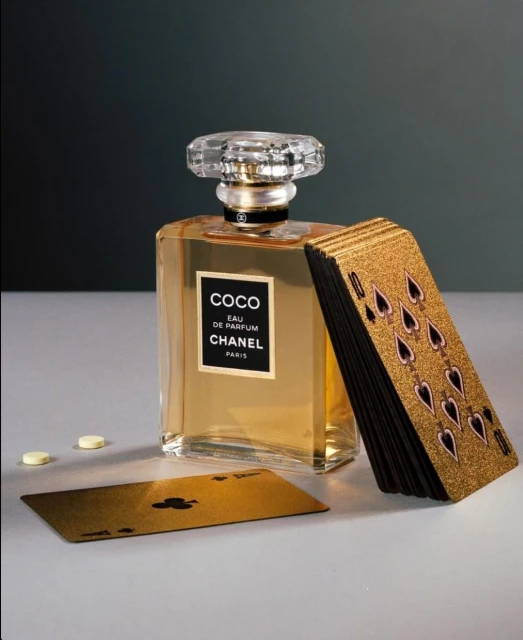 Elegant still life composition featuring Coco Chanel perfume bottle alongside a gold deck of playing cards and three pills on a white surface.