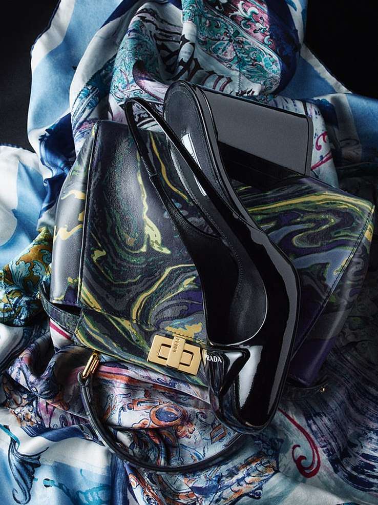 Fashion still life featuring a black patent leather purse and matching high heel shoes, set against a decorative scarf backdrop.