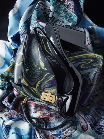 Fashion still life featuring a black patent leather purse and matching high heel shoes, set against a decorative scarf backdrop.