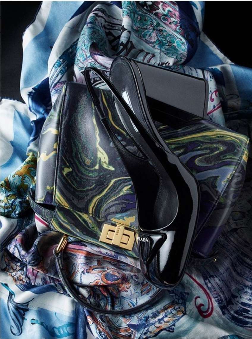 High-angle shot of a glossy black stiletto and a patterned Prada purse set against a backdrop of a swirling, colorful scarf.
