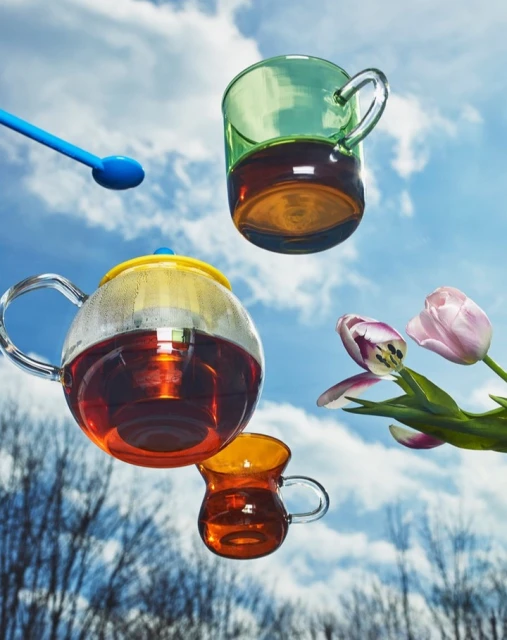 Still life of floating tea set with teapot, cups, blue spoon, and tulip flowers against a blue sky with cloudscape background.