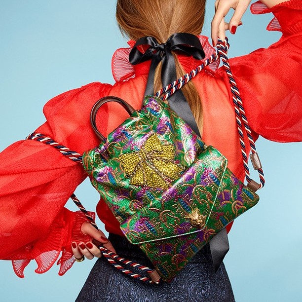 Woman with auburn hair poses from the back wearing a red top and patterned backpack with colorful rope straps against a blue background.