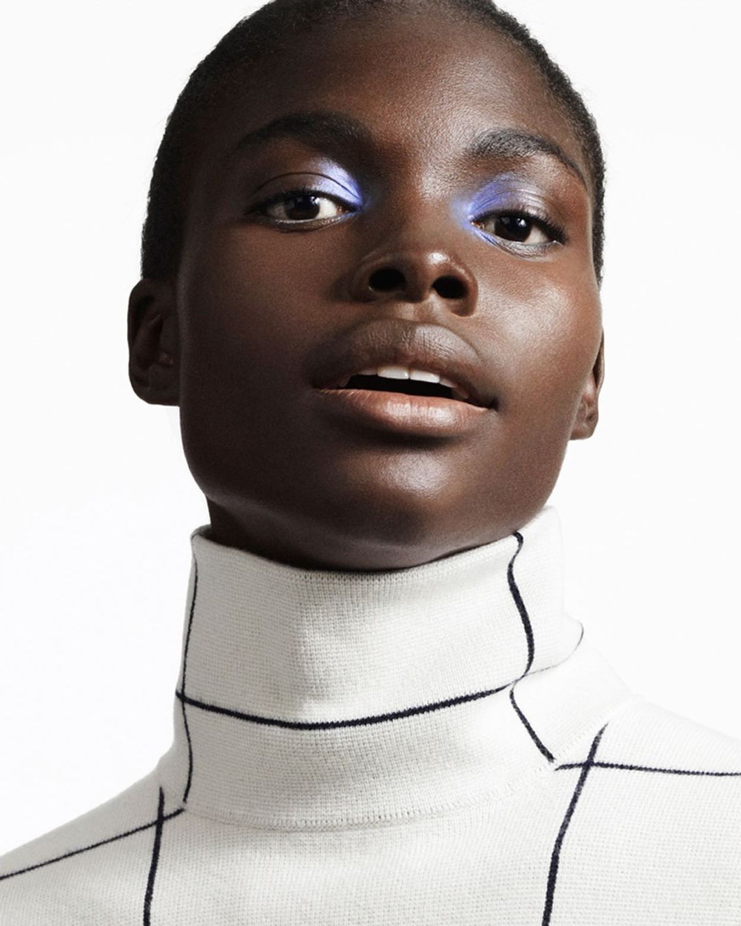 Close-up portrait of a Black woman with short hair wearing a patterned turtleneck sweater and bright blue eyeshadow against a white background.