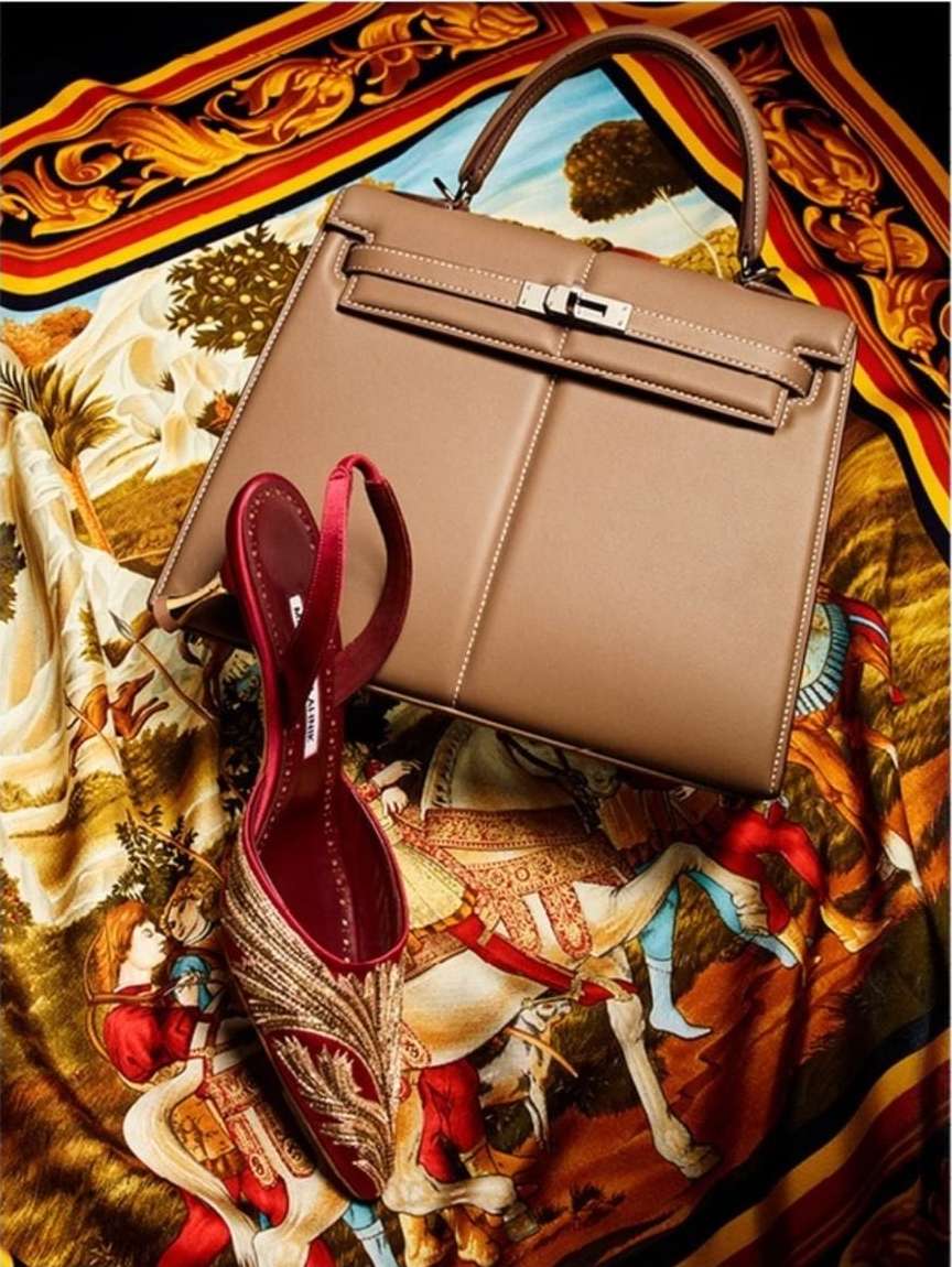 Fashion still life featuring a tan leather handbag and a red high-heeled shoe with gold embroidery on a printed fabric backdrop.