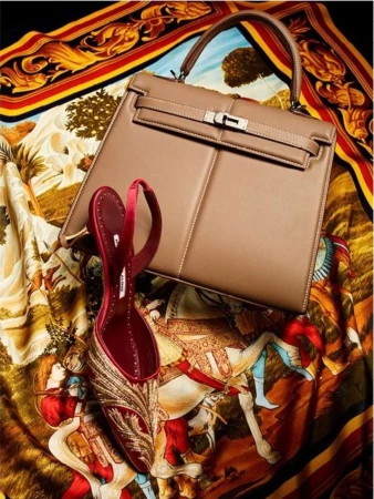 Fashion still life featuring a tan leather handbag and a red high-heeled shoe with gold embroidery on a printed fabric backdrop.