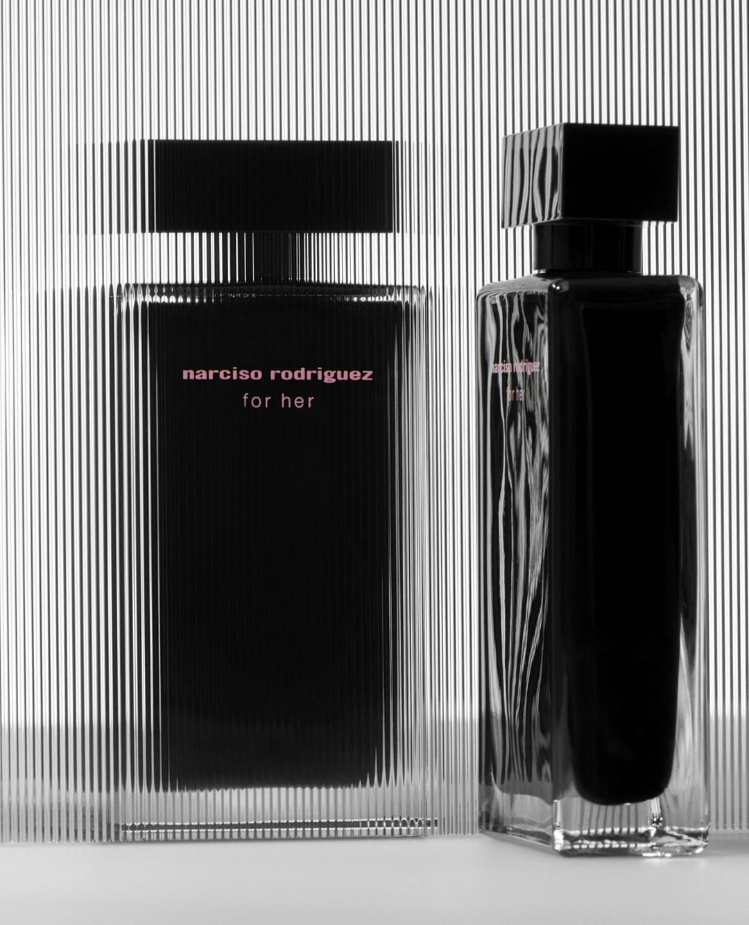 Two Narciso Rodriguez perfume bottles stand against a striped background, featuring a black and clear design with pink lettering.
