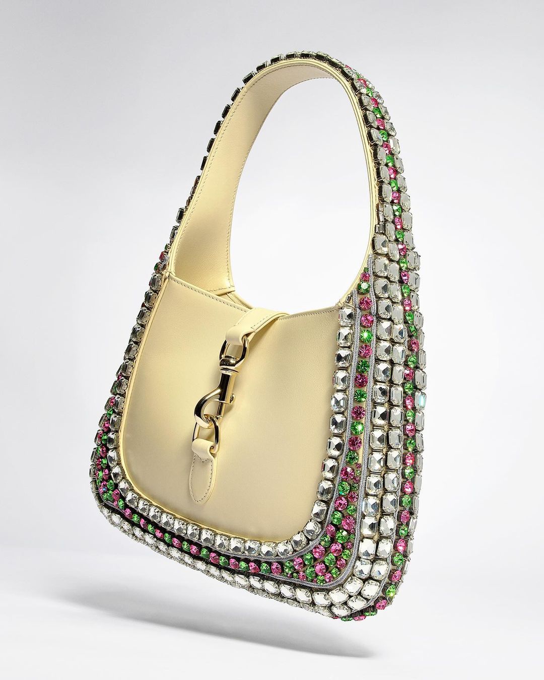 Luxury cream-colored leather handbag adorned with clear, pink, and green gemstones, featuring a silver clasp, in a studio product shot.