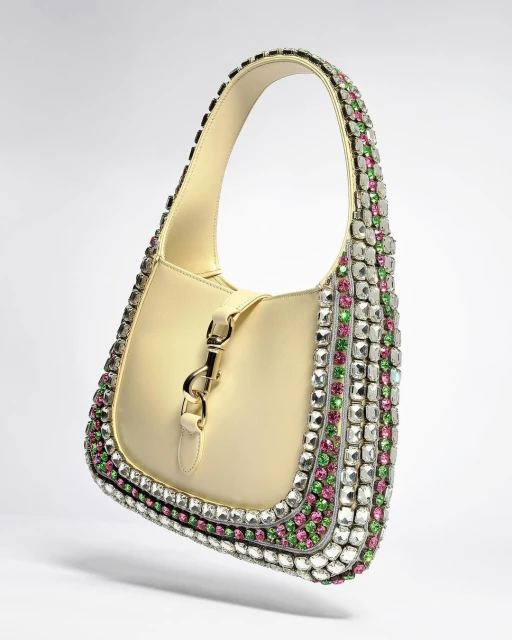 Luxury cream-colored leather handbag adorned with clear, pink, and green gemstones, featuring a silver clasp, in a studio product shot.