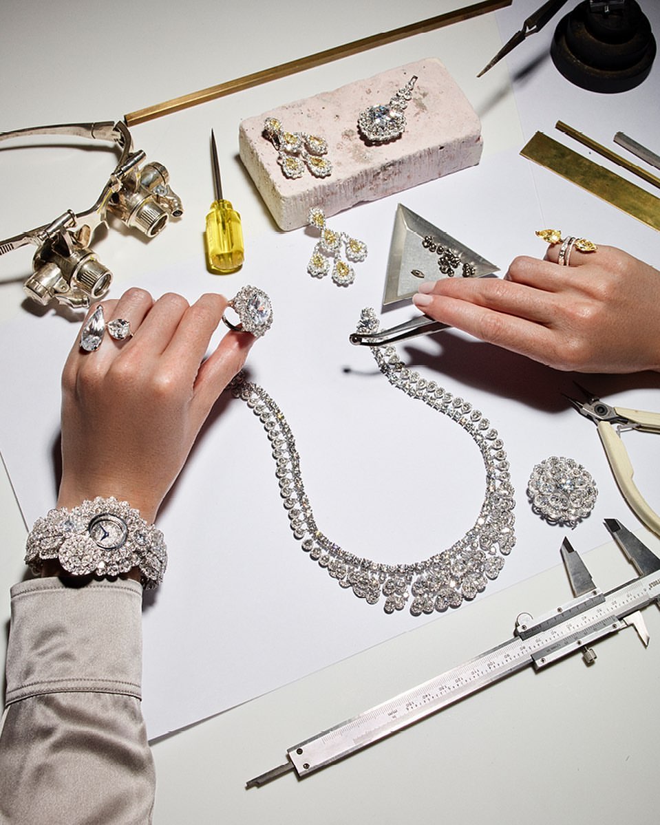 Close-up of jewelry arrangement with a wristwatch, necklace, earrings, and rings displayed on a white surface, with hands visible.