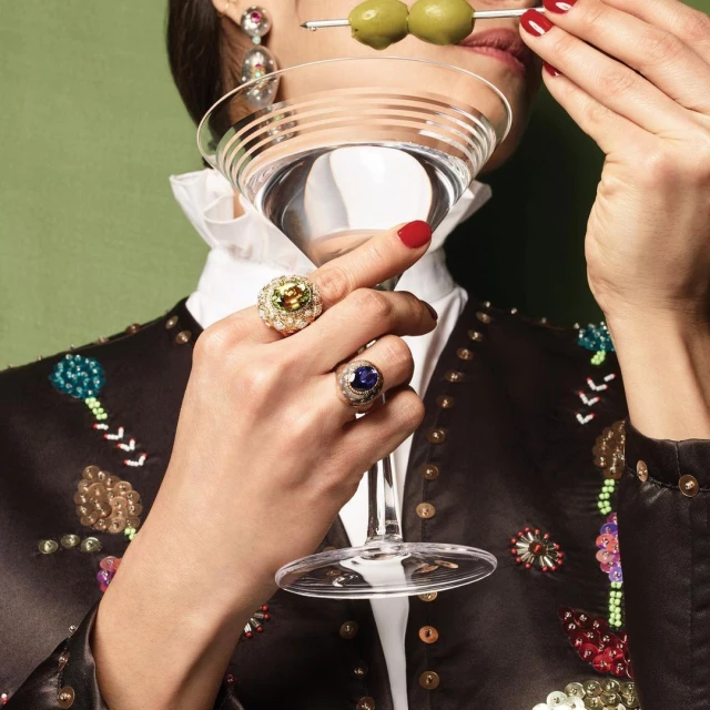 Woman holds martini with olives, showcasing jewelry. Rings on fingers, large earrings, black jacket, and red manicure against green.