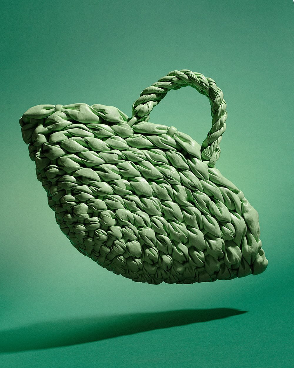 Elevated view of a woven green handbag floating in mid-air against a green background, casting a shadow on the seamless backdrop.