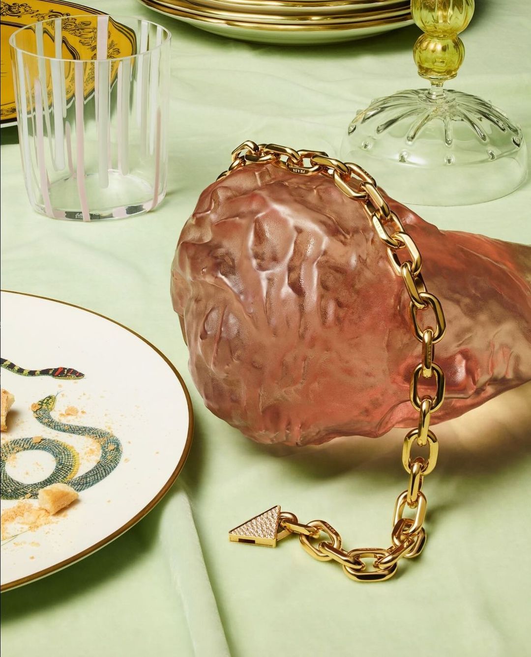 A gold chain link bracelet with a triangle clasp rests on a decorative pink stone, with tableware in the background.