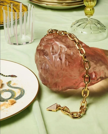 A gold chain link bracelet with a triangle clasp rests on a decorative pink stone, with tableware in the background.