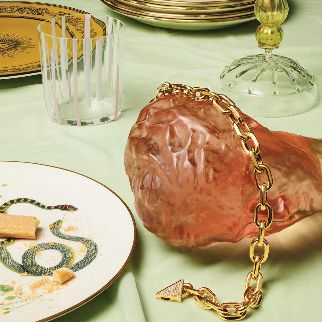Still life of a gold chain bracelet, clear glass, and a plate with a snake design, styled on a light green surface.