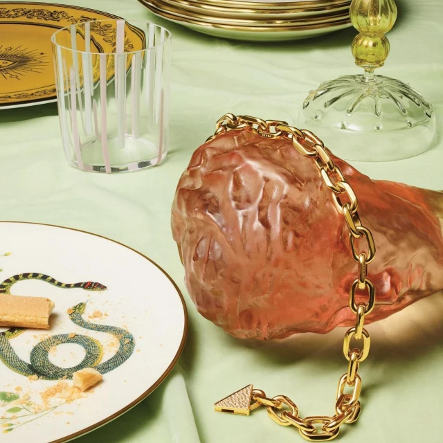 Still life of a gold chain bracelet, clear glass, and a plate with a snake design, styled on a light green surface.